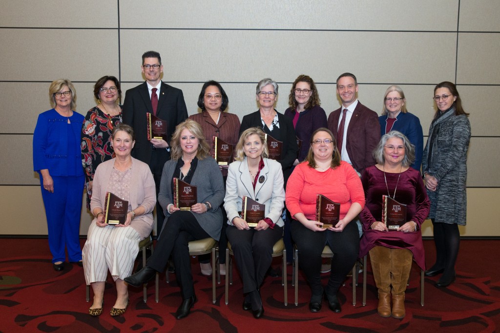 A group of 12 faculty members and 2 administrators, some sitting and some standing. I am sitting in a coral top and holding my award.